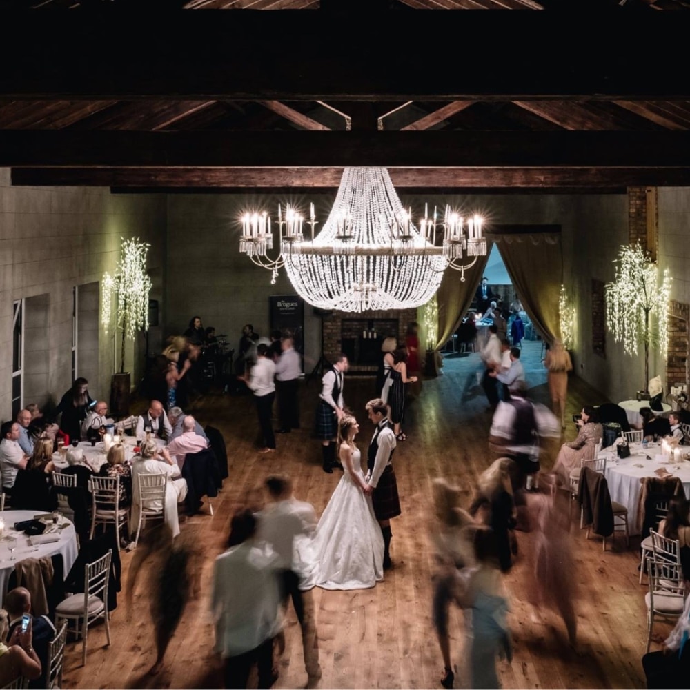 Elegant wedding reception in a warmly lit hall with a large chandelier, featuring a bride and groom dancing.