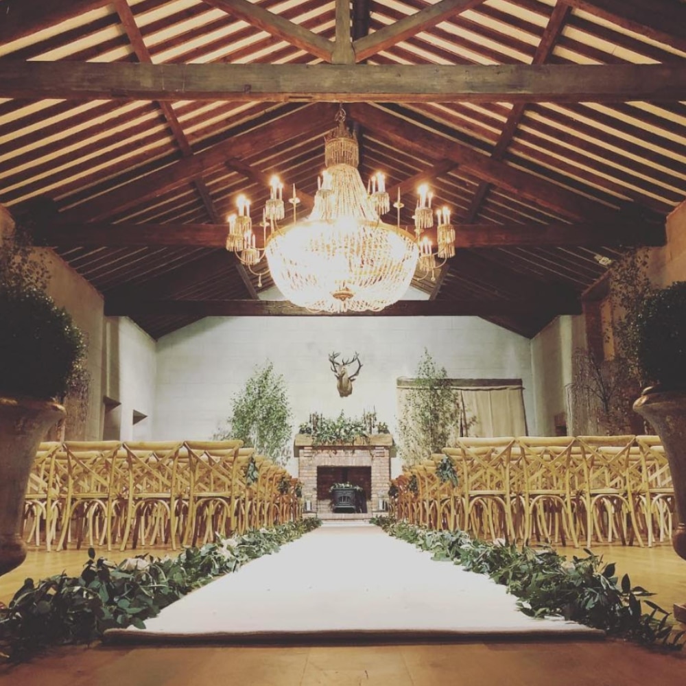 Elegant wedding venue with a grand chandelier, wooden chairs, and a decorated aisle leading to a stone fireplace.