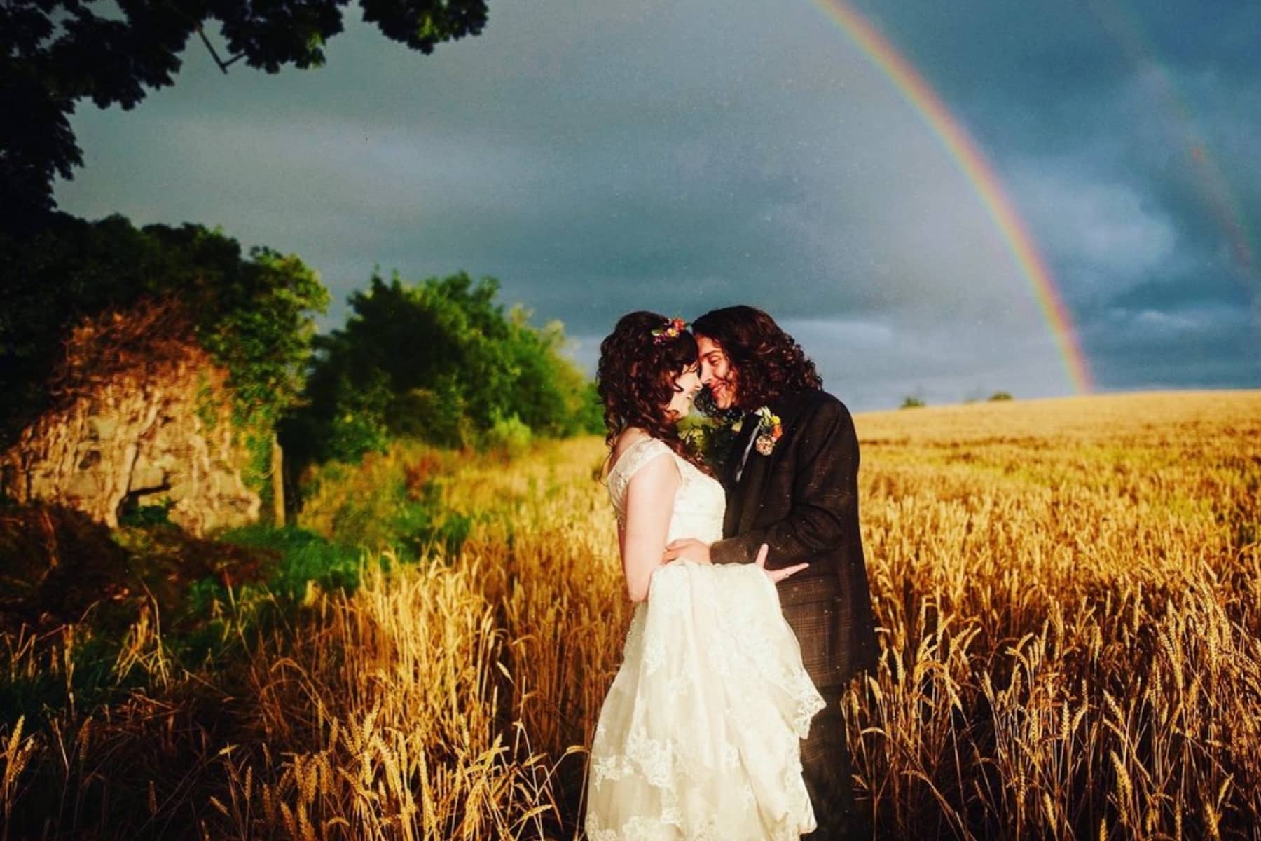 Bride and groom embracing in a wheat field under a vibrant rainbow.