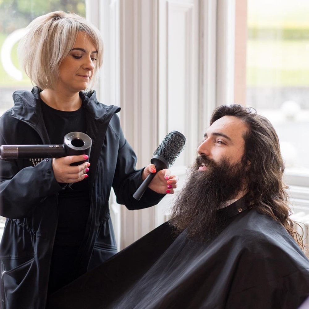Hairdresser styling a man's long hair and beard with a brush and blow dryer in a salon.