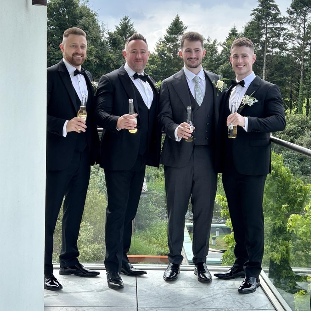 Four men in formal suits holding drinks on a balcony with trees in the background.