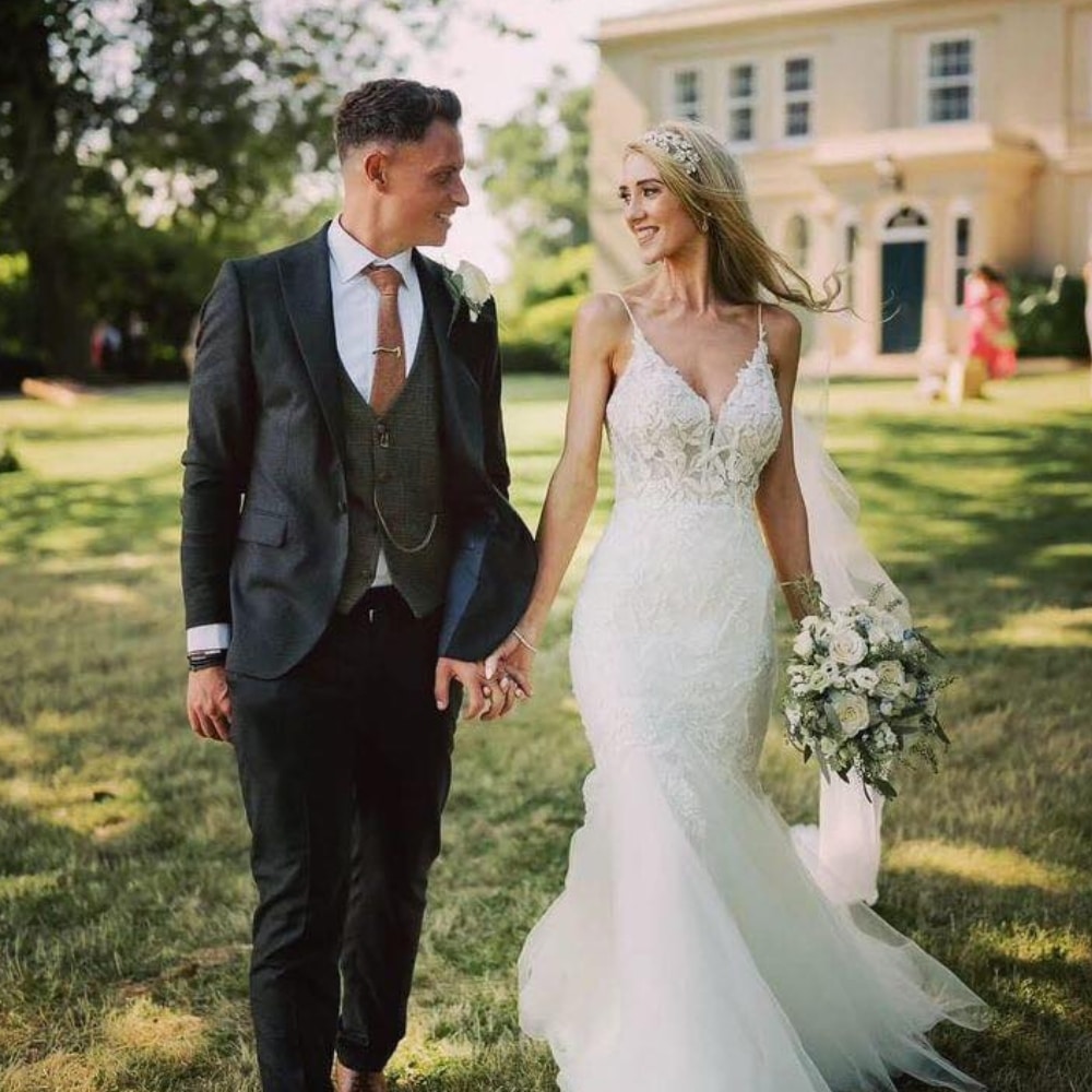Bride and groom holding hands in a garden, dressed in wedding attire, smiling at each other.