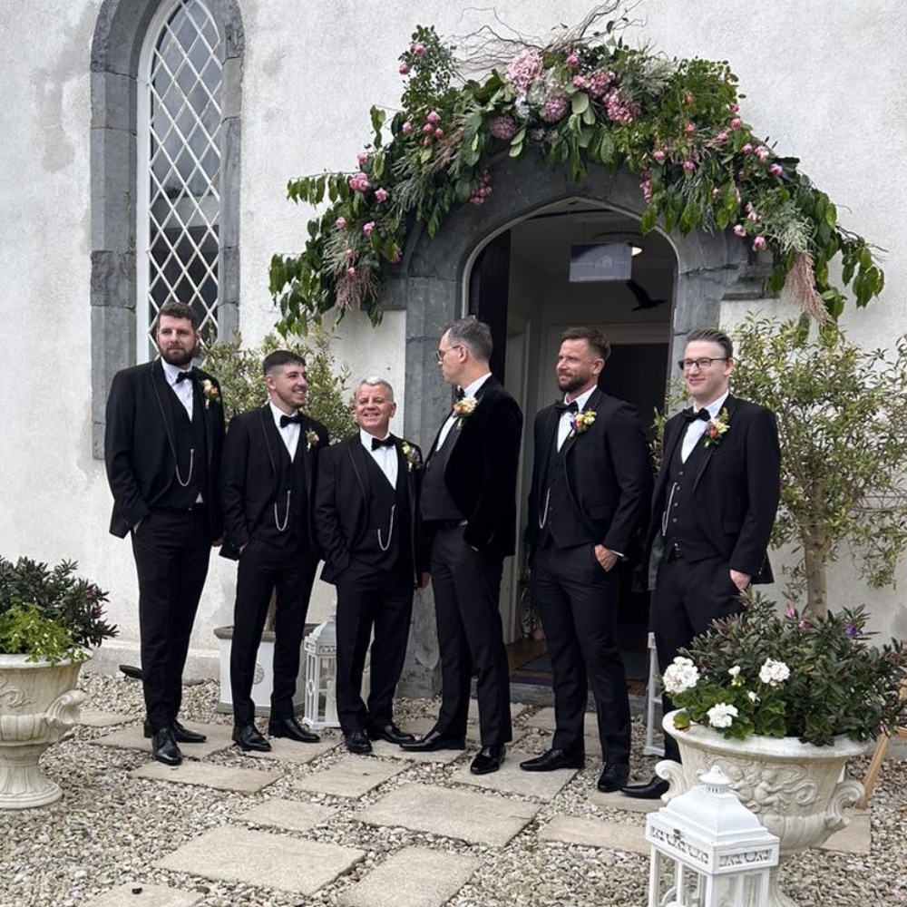 Groomsmen in tuxedos stand outside a decorated archway at a wedding venue.