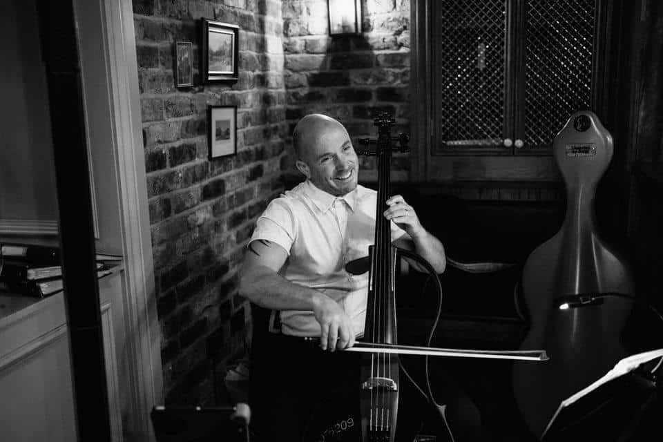 Musician playing a cello indoors, smiling, with a brick wall backdrop.