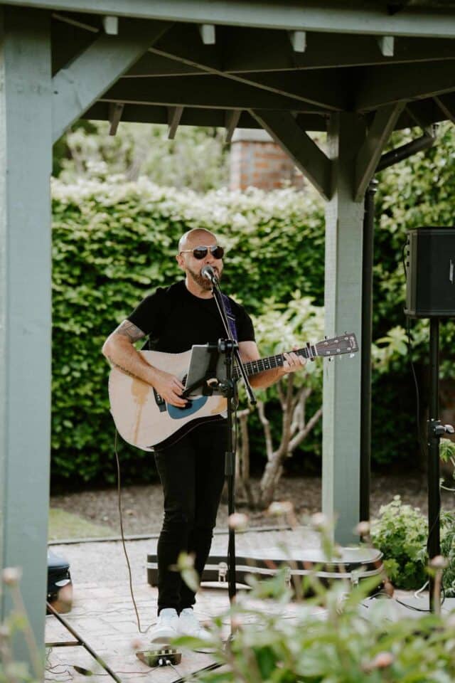 Musician playing guitar and singing under a gazebo outdoors.
