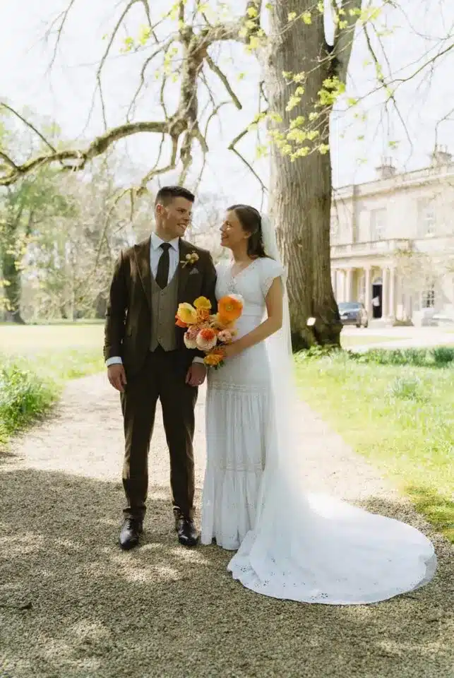 Bride and groom standing outdoors with bouquet under trees.