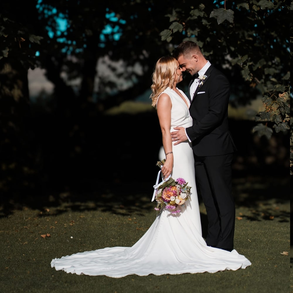 Bride and groom embracing outdoors, bride holding a bouquet, surrounded by greenery.