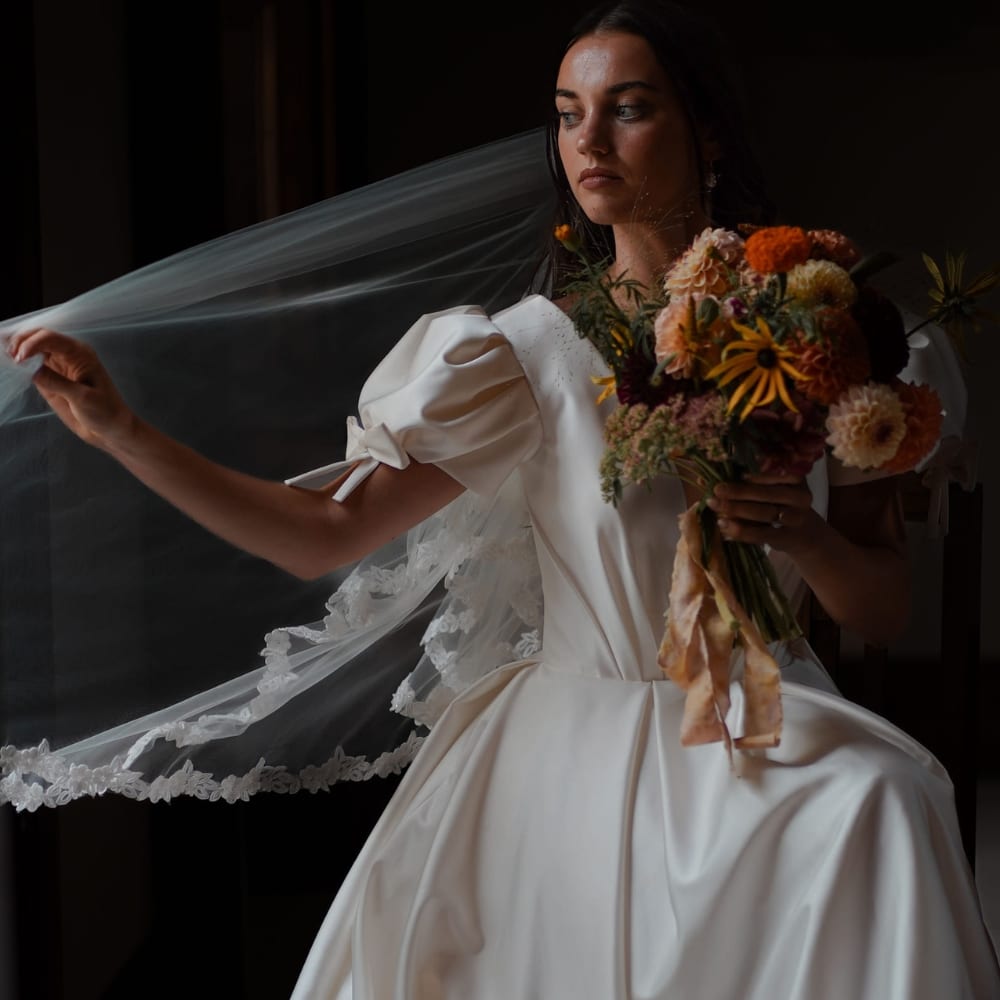 Bride in elegant gown holding bouquet, adjusting lace veil.