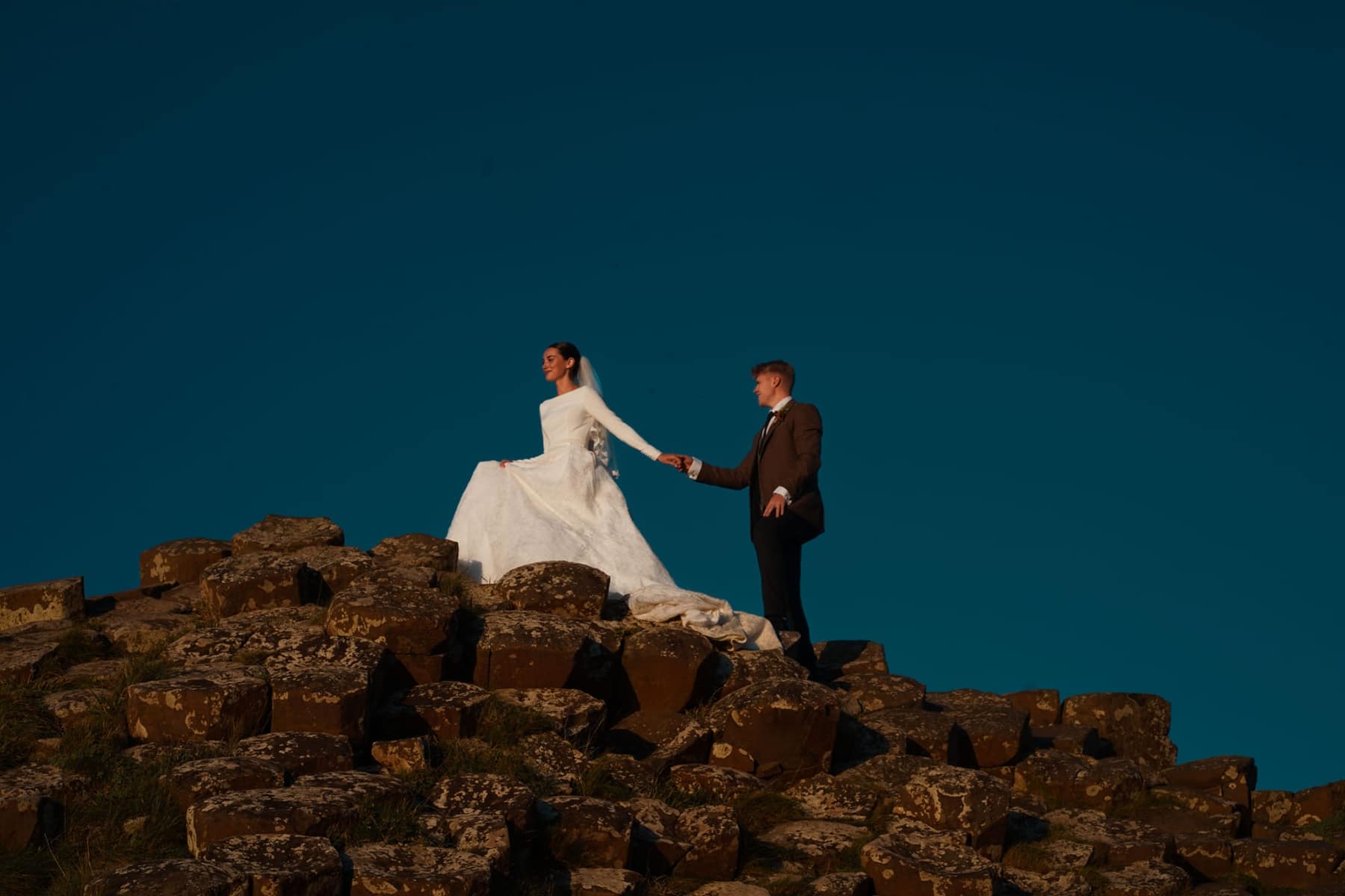 Bride and groom standing on rocky terrain under a clear blue sky, holding hands.