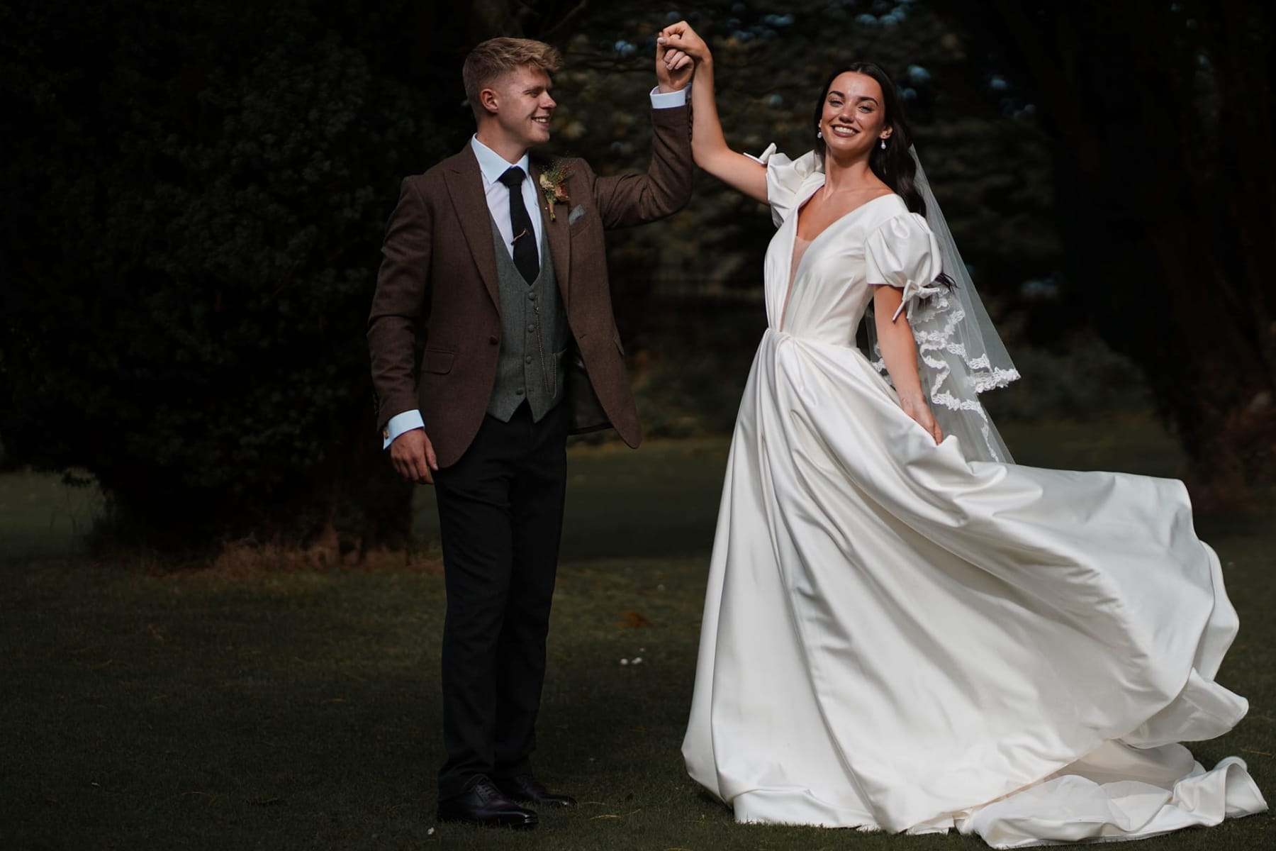 Bride and groom dancing in a garden, bride wearing white gown and veil, groom in brown suit.