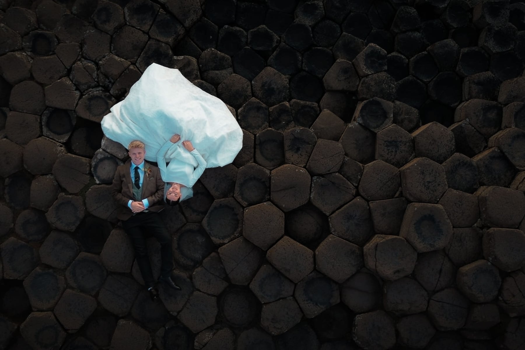 Aerial view of a couple in formal attire lying on hexagonal basalt columns, resembling a wedding photo shoot.