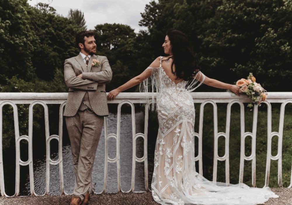 Bride and groom on a bridge, bride holding bouquet, groom leaning on railing, lush greenery in background.