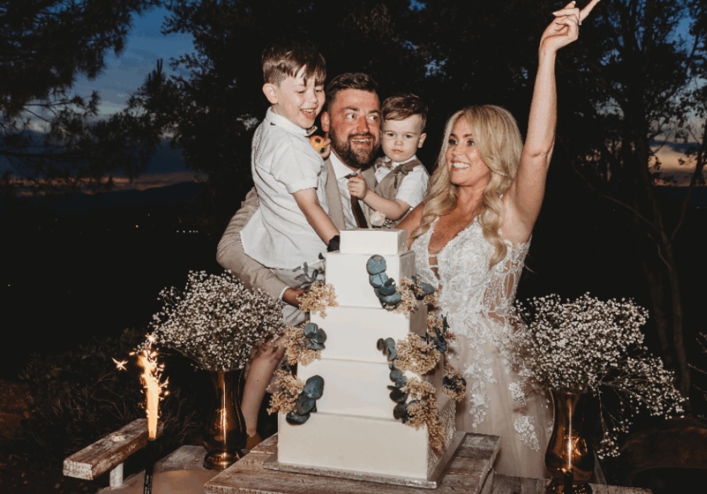 Family celebrating with a tiered white cake adorned with flowers outside at dusk.
