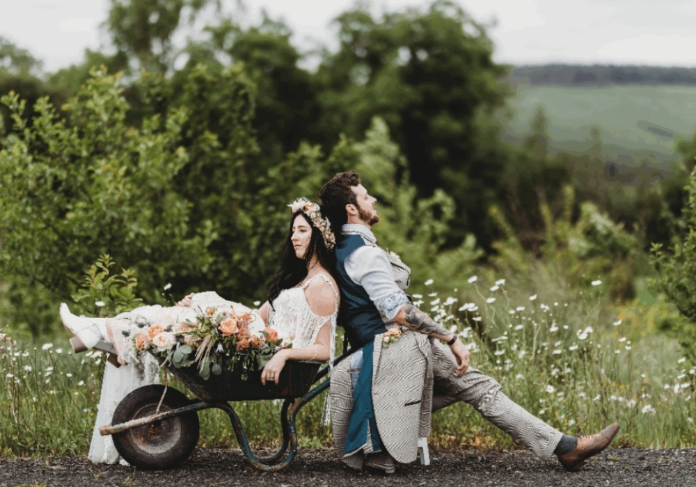 Bride in a wheelbarrow with flowers, groom seated back-to-back outdoors.