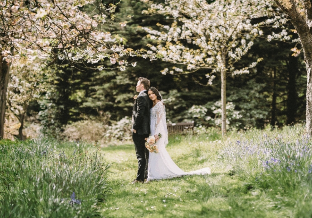 Bride and groom standing together in a lush garden with blooming trees and green grass.