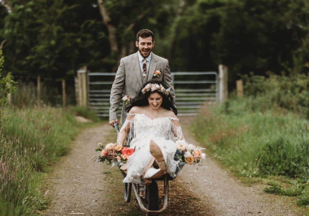 Bride in a wheelbarrow with flowers, pushed by groom on a rustic path.
