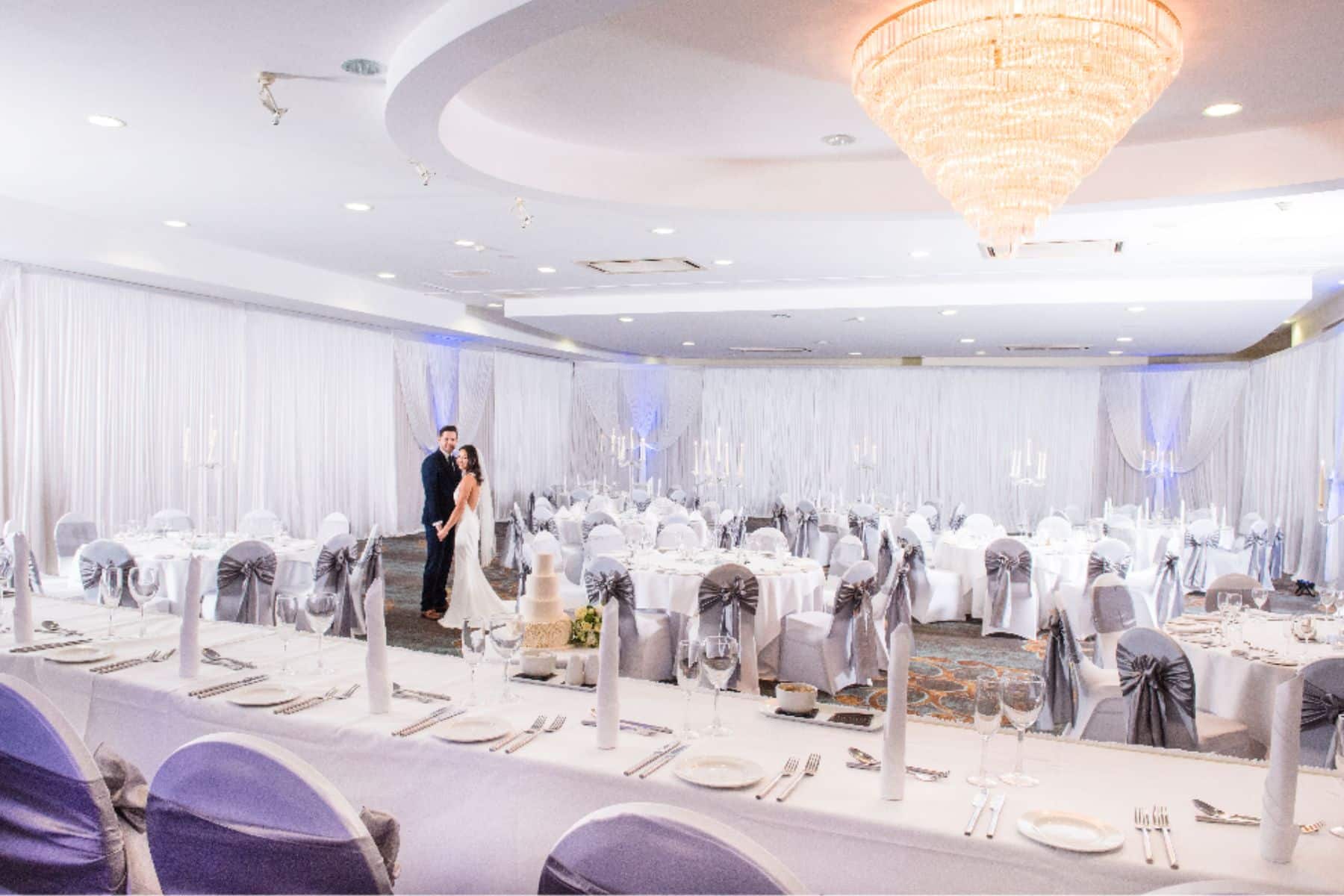 Elegant wedding reception setup with a couple in a decorated ballroom, featuring round tables, white linens, and a chandelier.