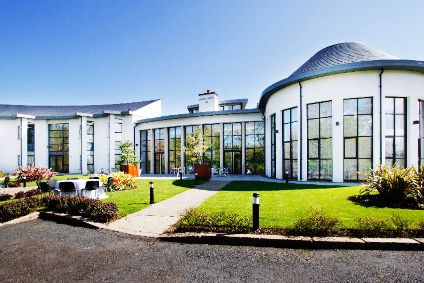 Modern hotel exterior with large windows, green lawn, and outdoor seating on a sunny day.