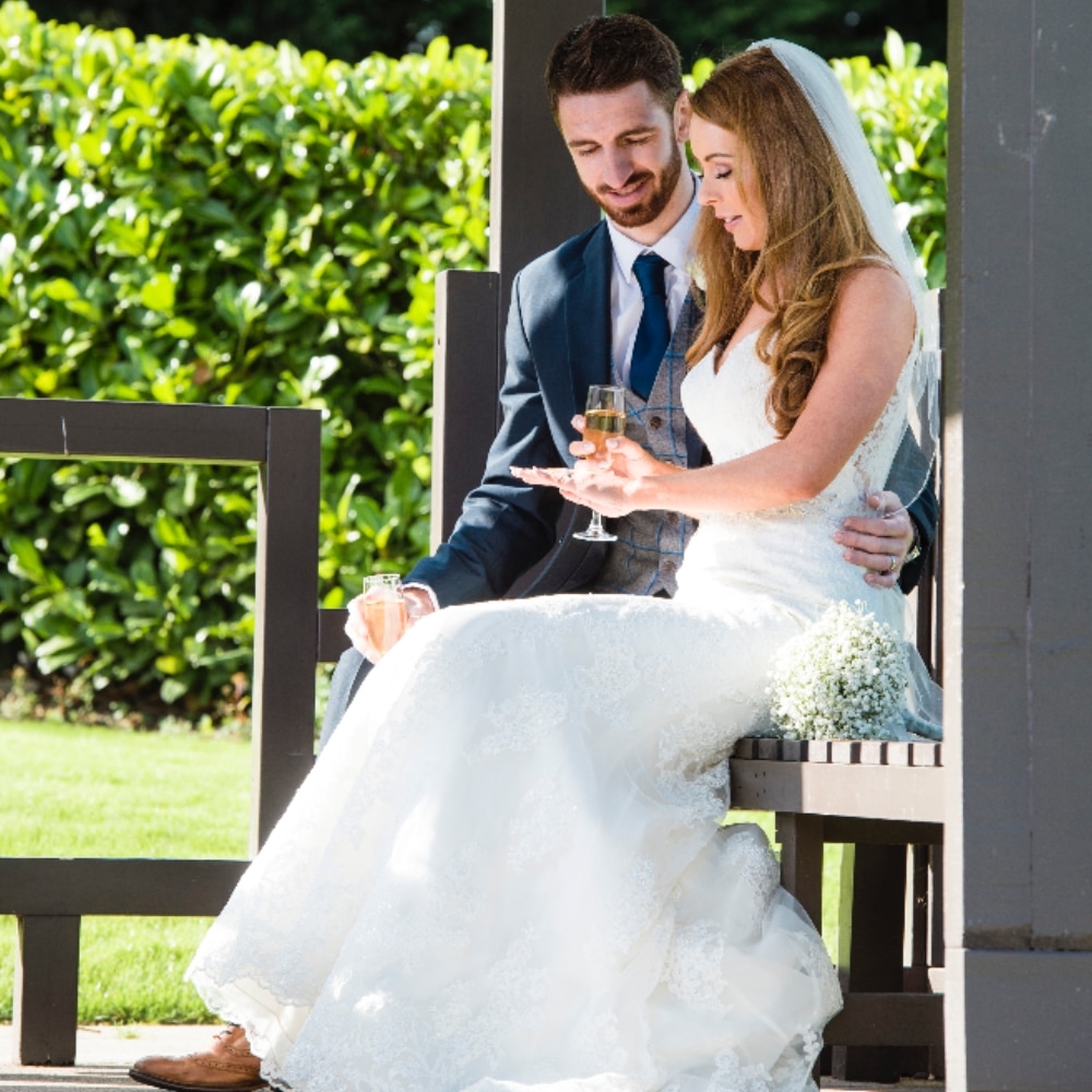 A bride and groom sit together, holding drinks, in a garden setting at La Mon Hotel.