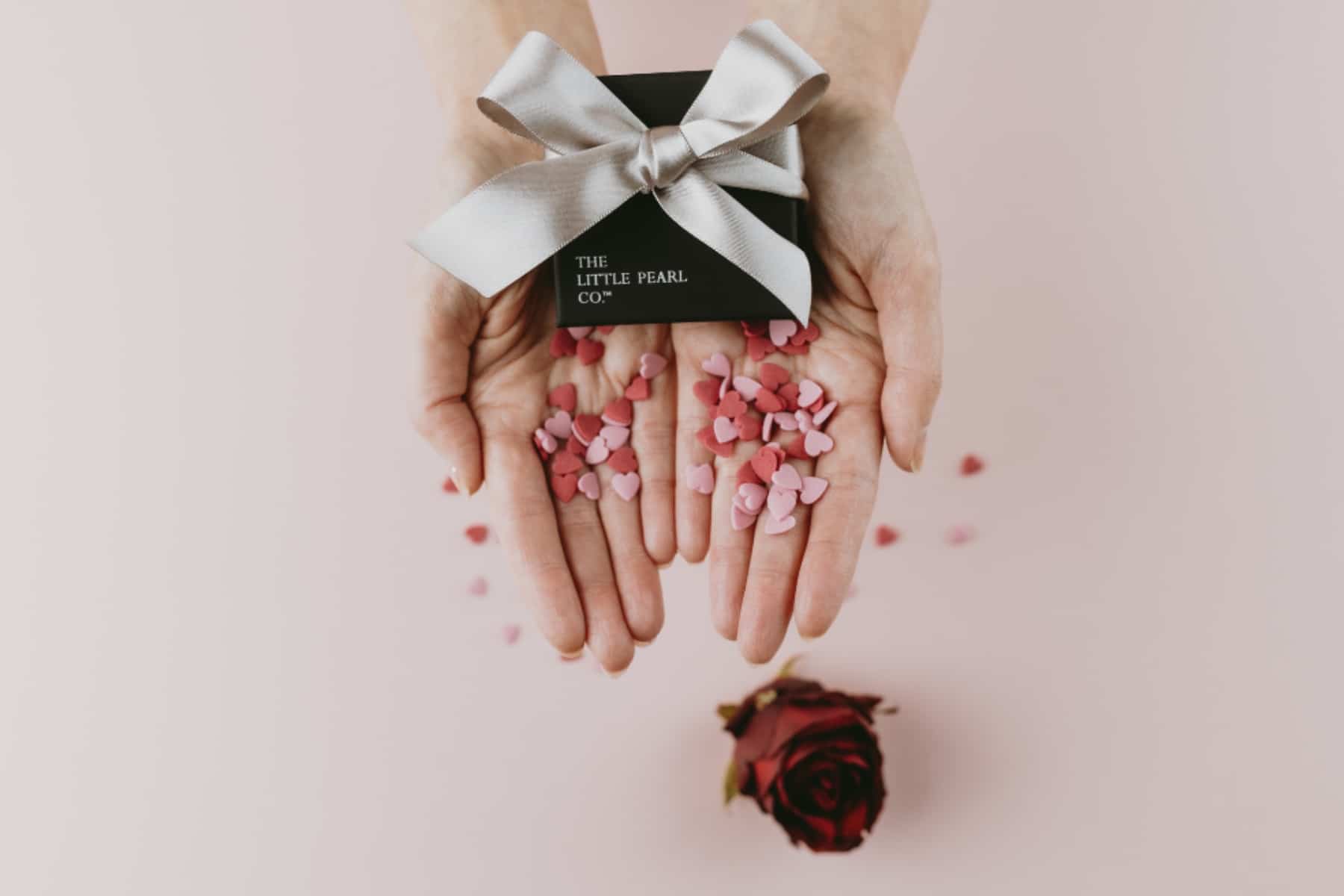 Hands holding a gift box tied with a silver ribbon, surrounded by pink heart-shaped confetti, with a red rose below.