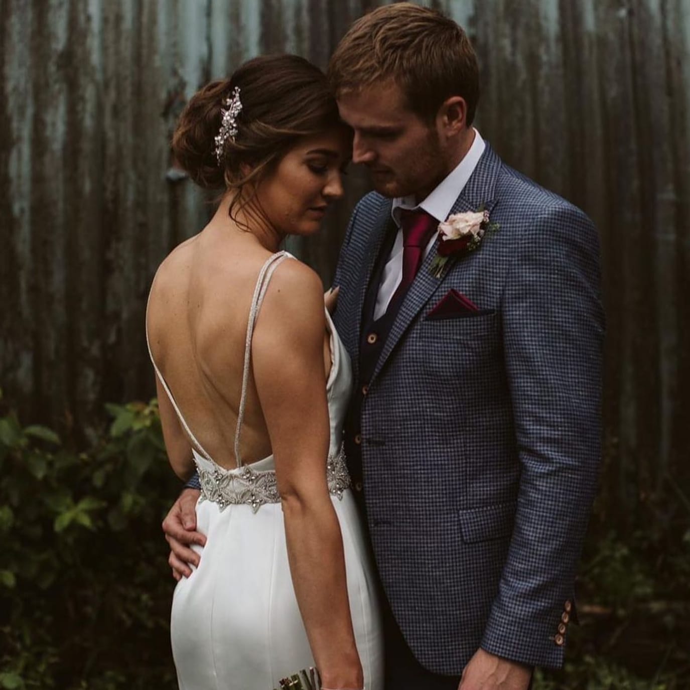 Bride and groom embracing, bride in white dress, groom in blue suit with red tie, outdoor setting.