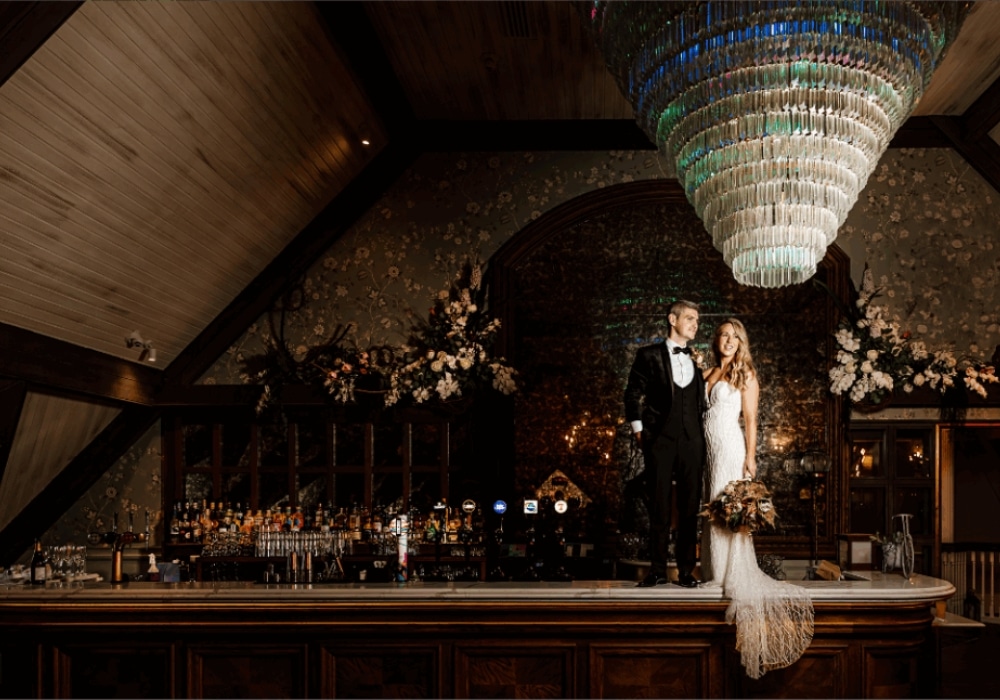 Couple standing on a bar counter in formal attire under a large chandelier.
