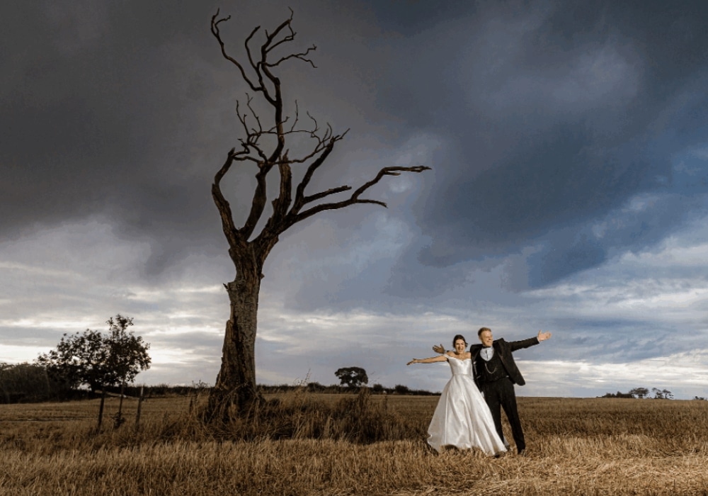 Bride and groom in wedding attire posing beside a barren tree under a cloudy sky.