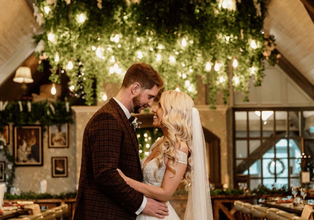 Bride and groom embracing under a canopy of lights and greenery in a warmly lit venue.