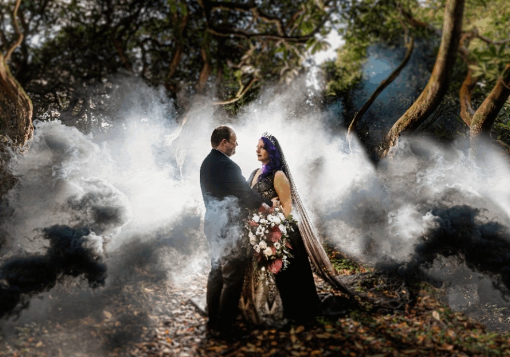 Bride and groom in a forest surrounded by mist, wearing formal attire with a bouquet of flowers.