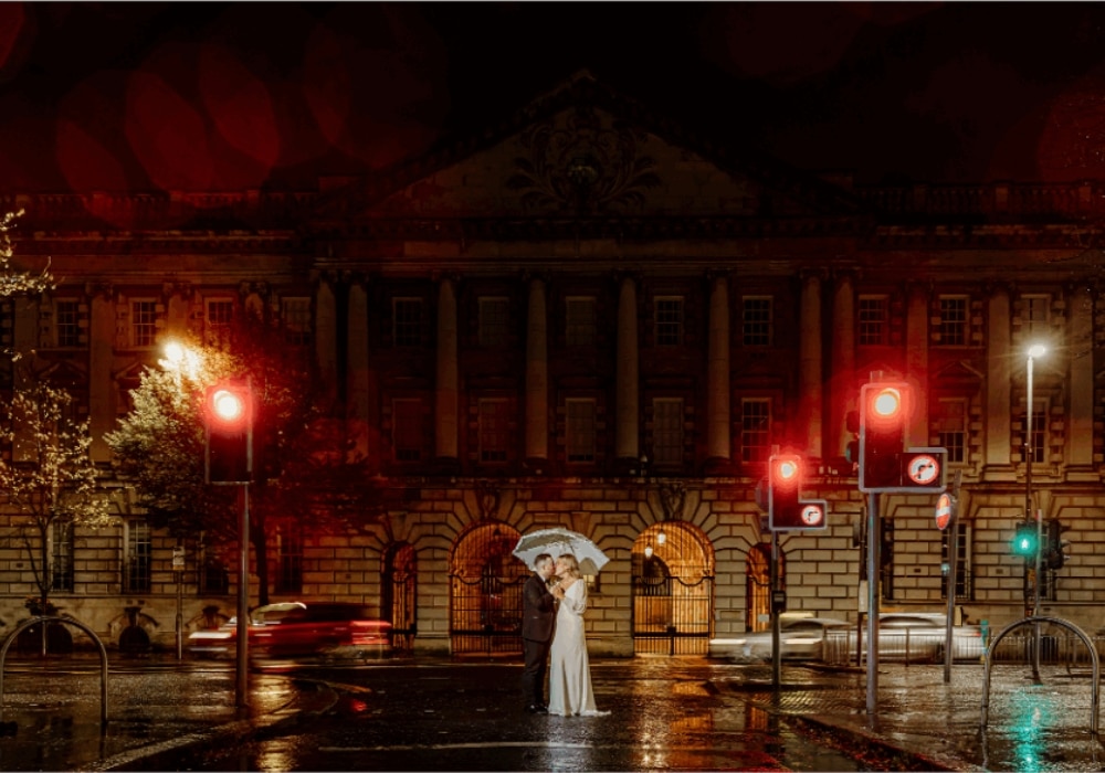 Couple under umbrella at night on wet street, surrounded by traffic lights and historic building.