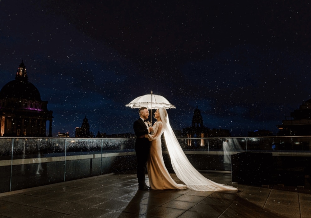 Bride and groom embracing under an umbrella at night, with city skyline and raindrops visible.