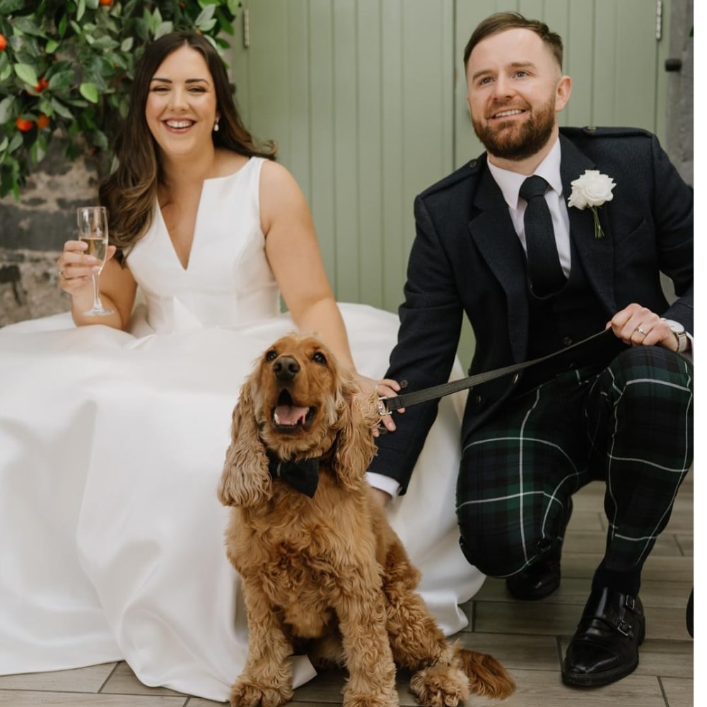 Bride and groom smiling with their dog wearing a bow tie at a wedding.