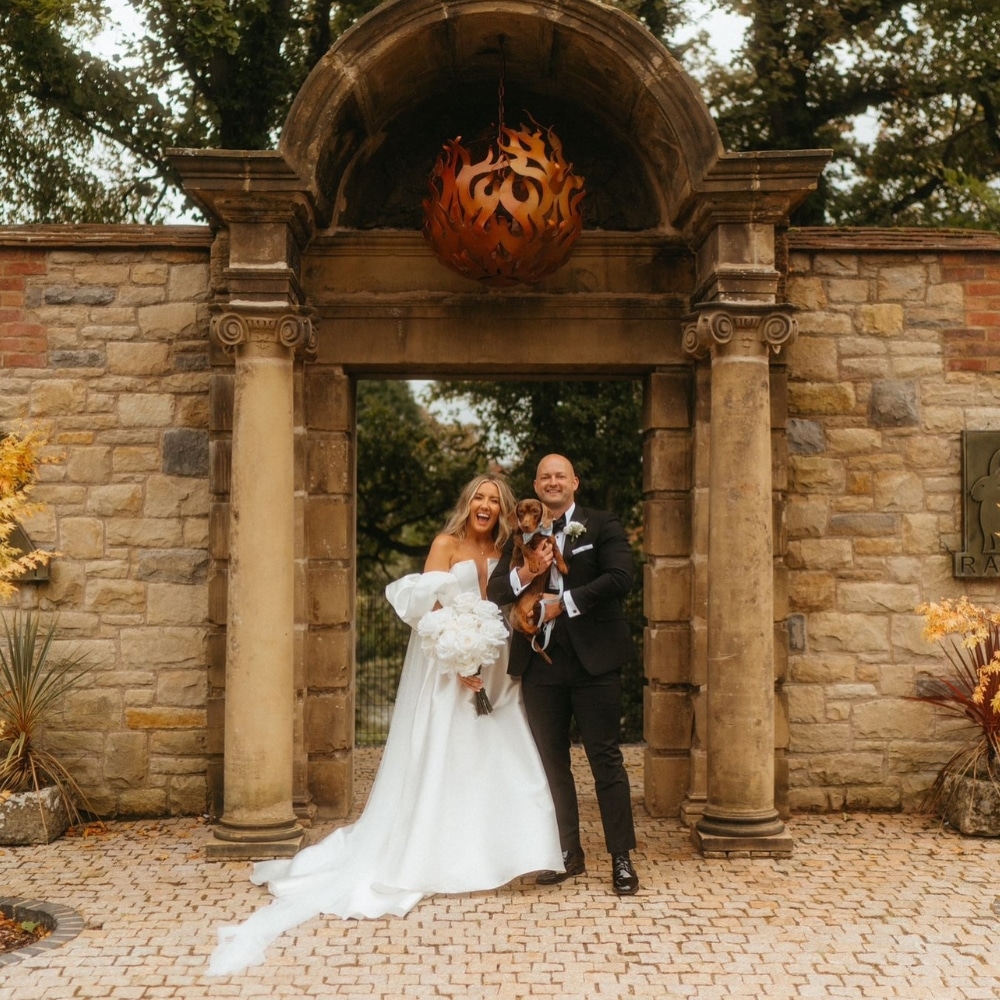 Bride and groom smiling, holding a dog, standing in front of a stone archway.