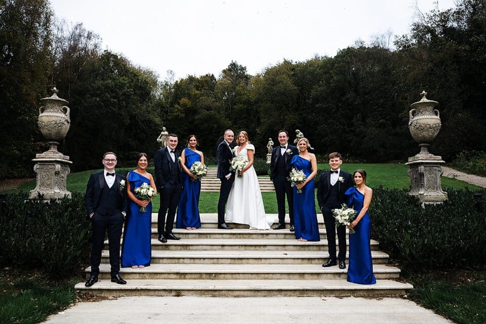 Wedding party on outdoor steps, with bride and groom surrounded by bridesmaids in blue dresses and groomsmen in suits.