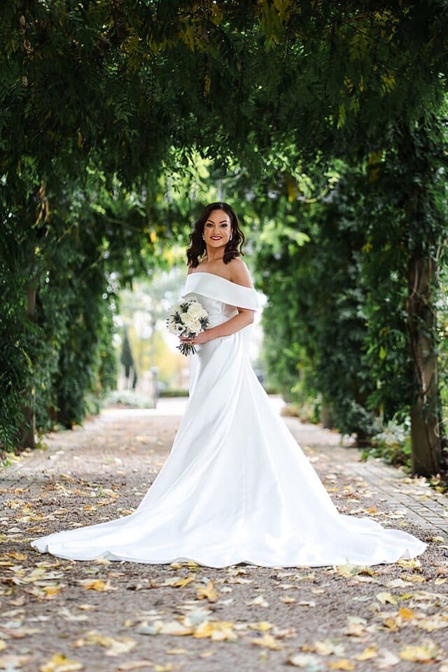 Bride in white off-shoulder gown holding bouquet, standing under green archway.