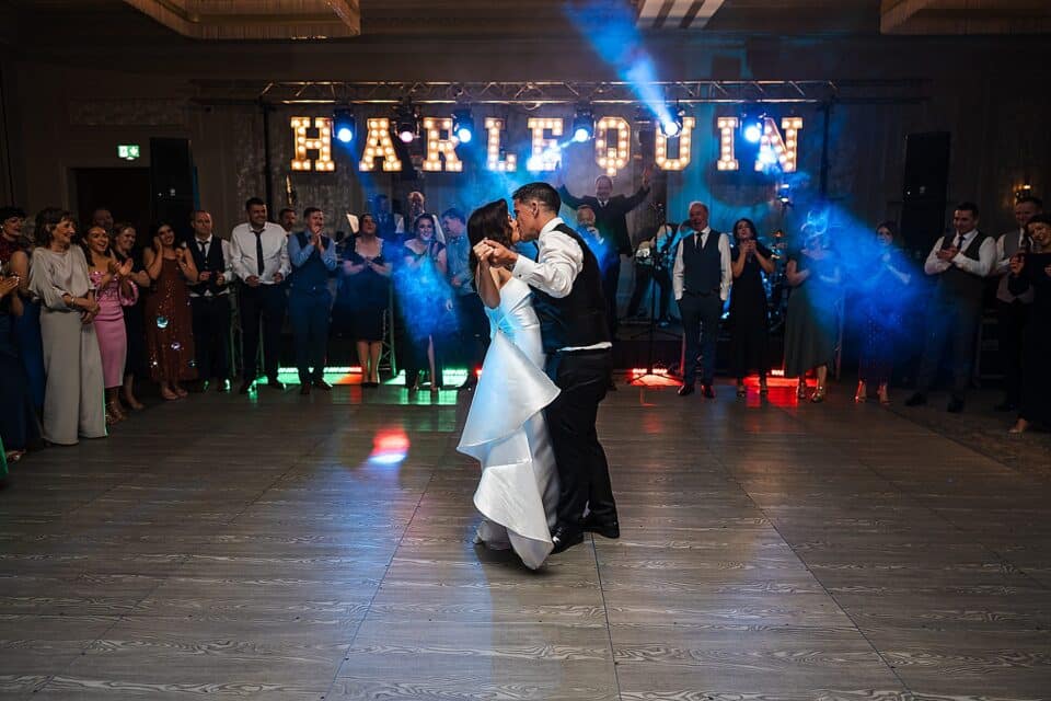 Bride and groom dancing at wedding with guests clapping and blue stage lights.
