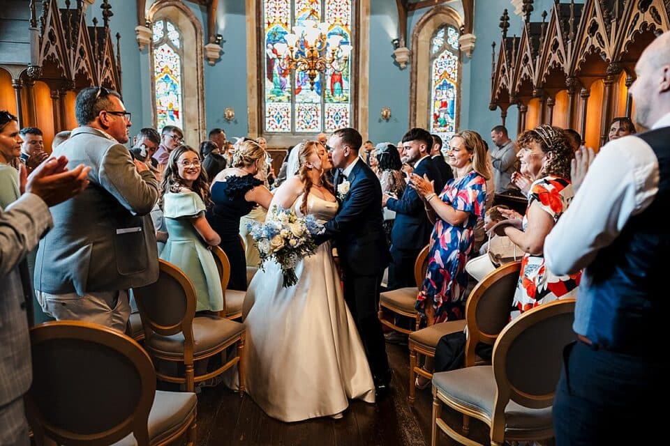 Bride and groom kiss amidst applauding guests in a church with stained glass windows.