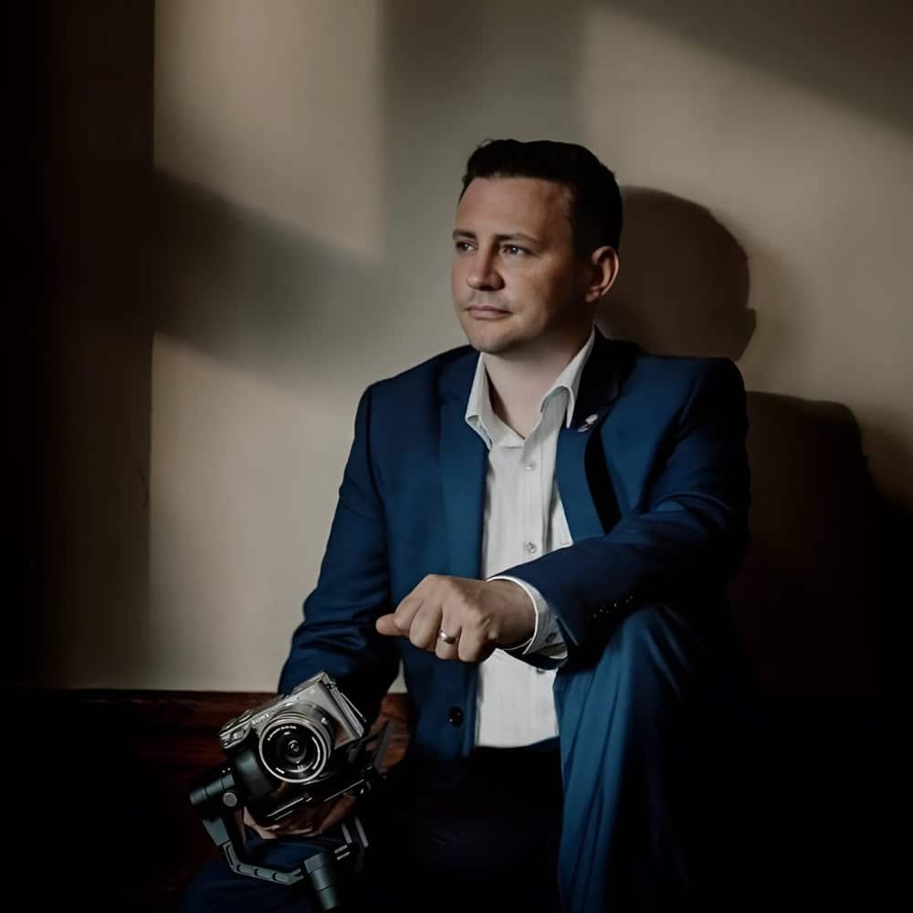 Man in a blue suit holding a camera, sitting in soft lighting.