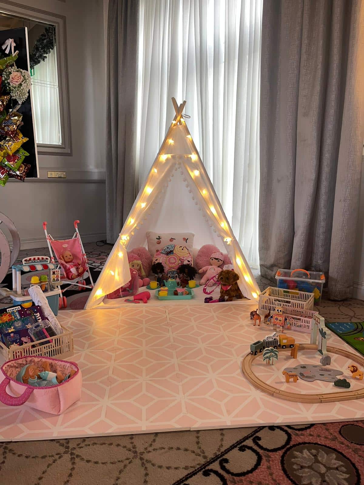 Children's playroom with a lit teepee tent, toys, and stuffed animals on a patterned mat.