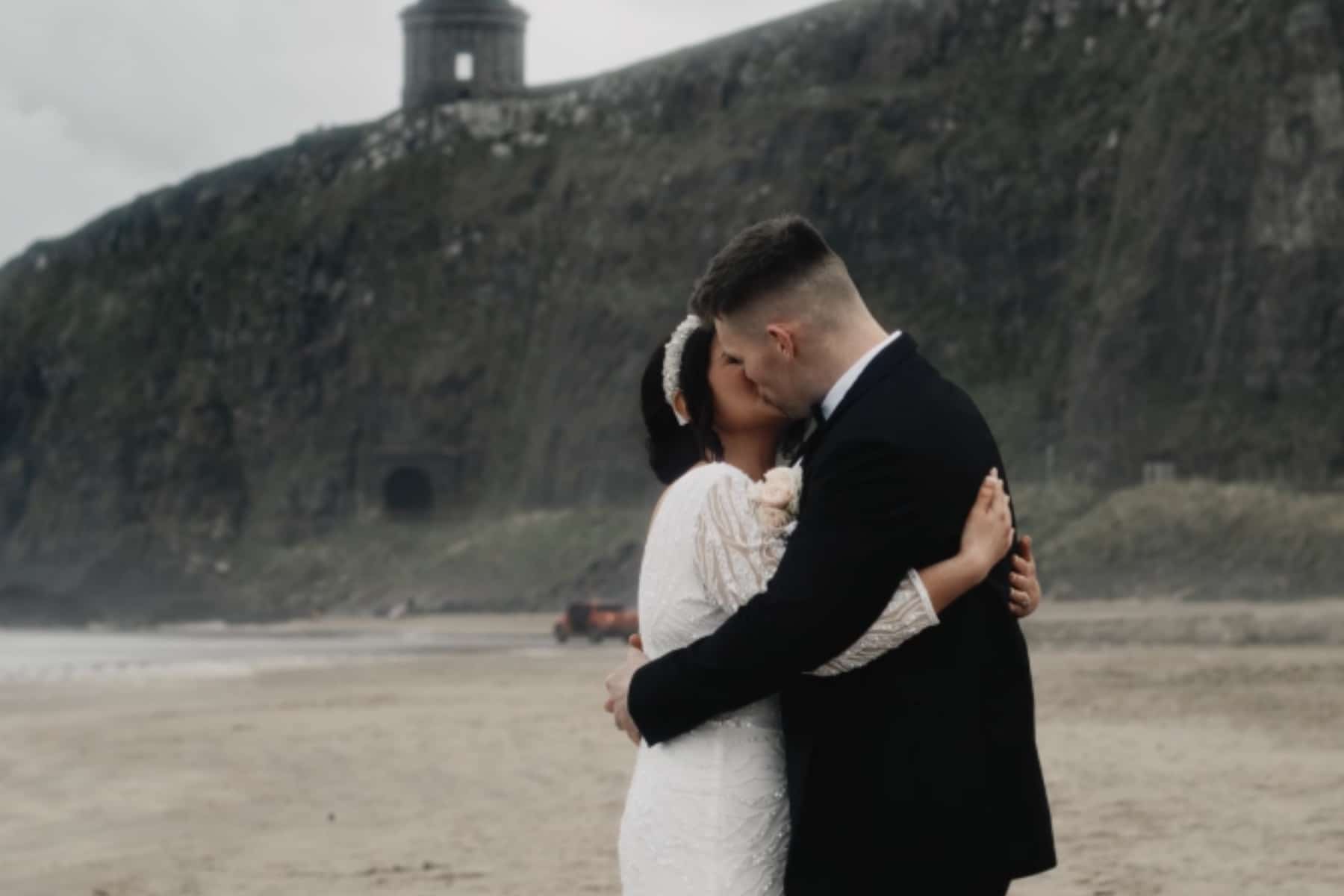 Bride and groom kissing on a beach with a rocky cliff and tower in the background.