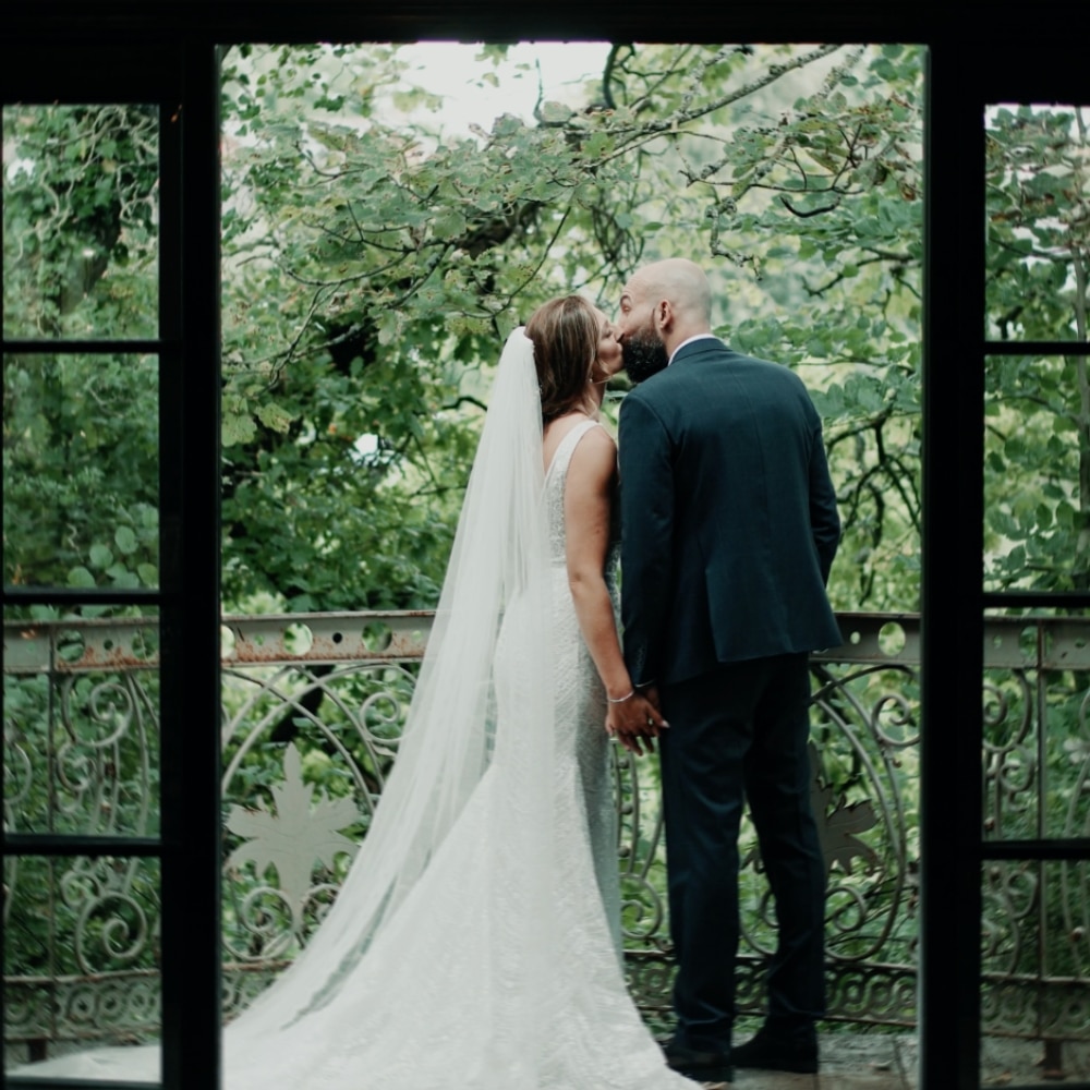 Bride and groom share a kiss on a balcony surrounded by lush greenery.