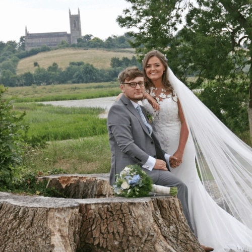 Bride and groom pose by a tree stump, with a scenic view of fields and a distant church on a hill.