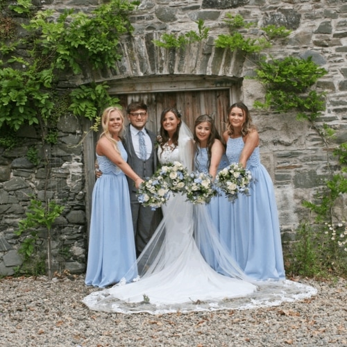 Bride and bridesmaids in blue dresses posing with bouquets in front of a rustic stone wall.