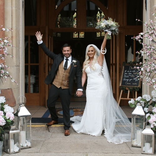 Bride and groom celebrate outside a decorated venue with lanterns and flowers, holding hands and waving enthusiastically.