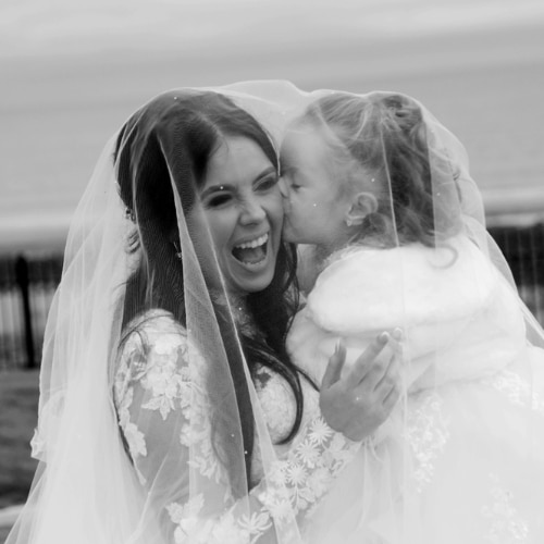 Bride laughing receiving a kiss from a young girl outdoors.