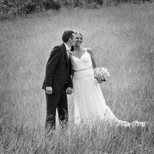 Bride and groom in a field, groom kissing bride's cheek, black and white photo.