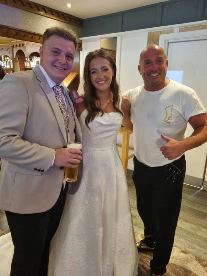 Three people smiling, one in a beige suit holding a drink, one in a white wedding dress, and another in casual attire indoors.