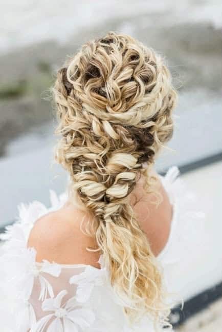 Intricate bridal hairstyle with blonde curls and braids, outdoor setting.
