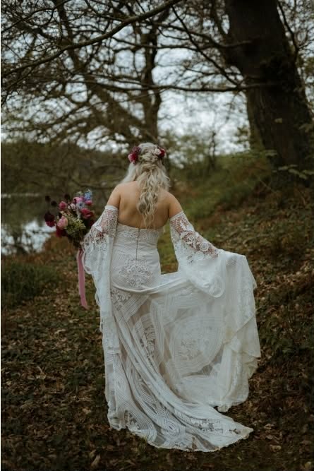 Bride in lace gown holding bouquet, walking through wooded area.