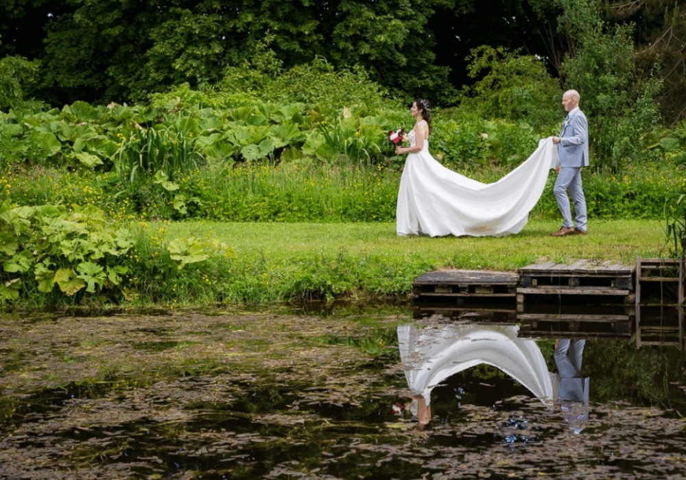 Bride and groom walking by a pond with greenery, the bride holding flowers.